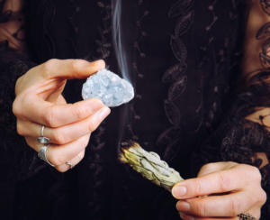 woman cleansing blue celestite crystal cluster gemstone by smudging white sage bundle.